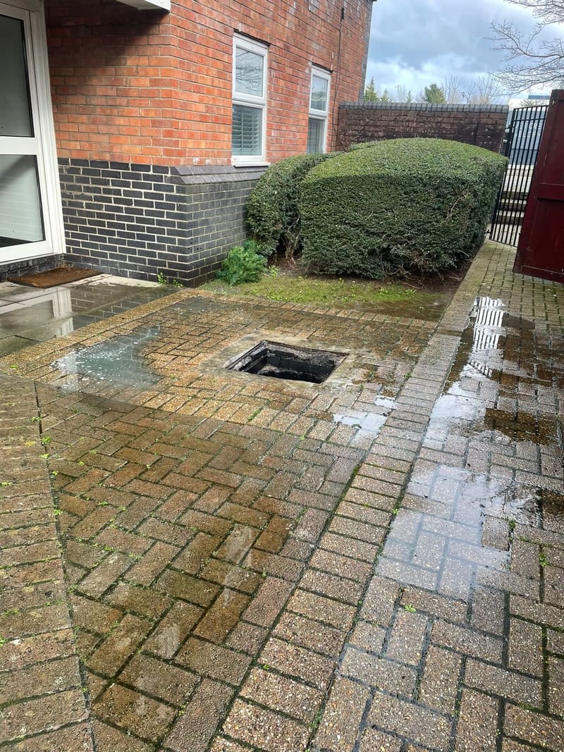 Open manhole in a wet brick-paved courtyard during drain jetting