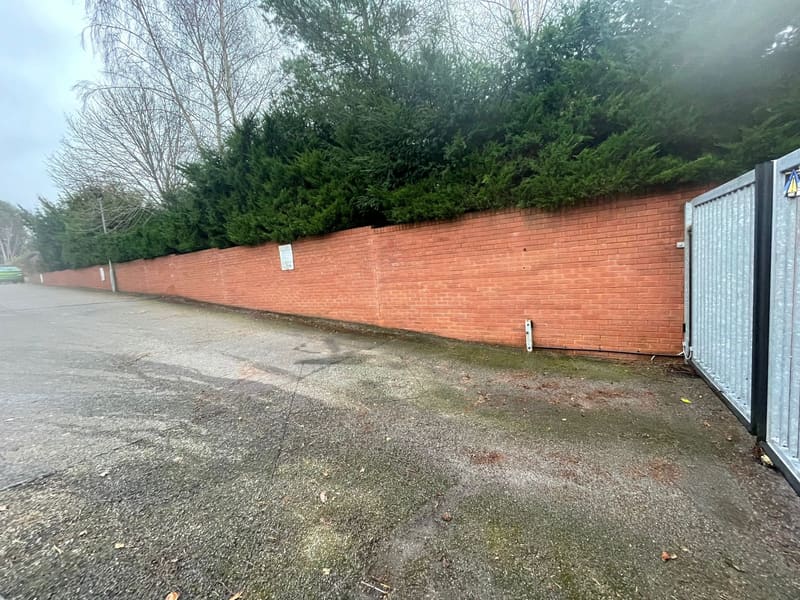 Long red brick wall with a hedge on top alongside a paved driveway
